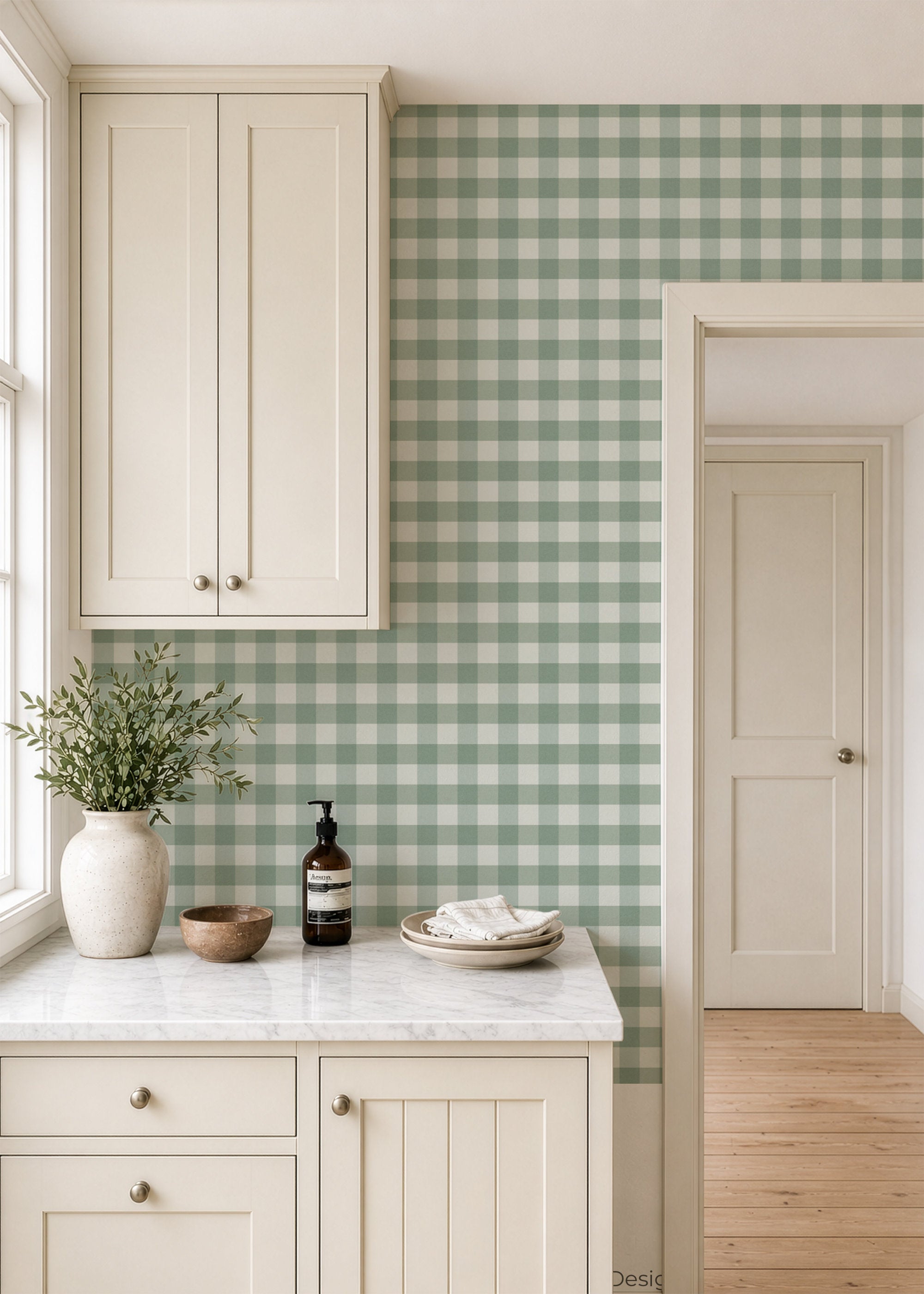 A styled kitchen interior featuring sage green and white gingham wallpaper on the backsplash, paired with cream-colored cabinets, a white marble countertop, and a vase of greenery.