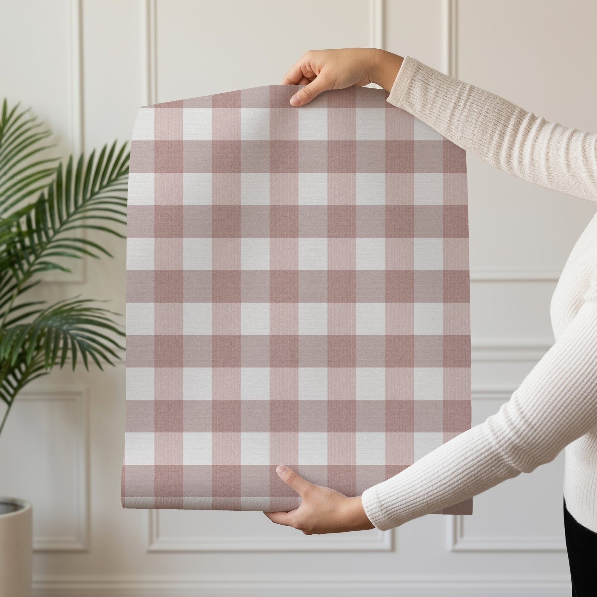 A person holds up a large sheet of pink gingham wallpaper featuring a dusty rose and white gingham check pattern, set against a neutral paneled wall with a potted plant in the background.